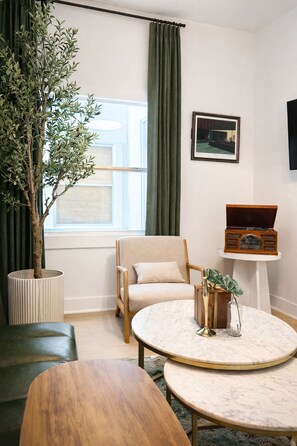 A modern living room with a wooden coffee table, a beige armchair, a potted plant, and a vintage radio on a white pedestal. - Newly renovated duplex conveniently located between Vilano and downtown. (St. Augustine)