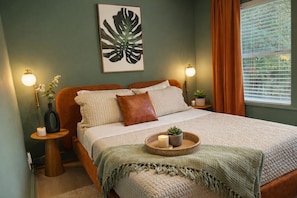 A bedroom with a bed, bedside tables, a wall-mounted light, a window with curtains, and a framed plant on the wall. - Newly renovated duplex conveniently located between Vilano and downtown. (St. Augustine)