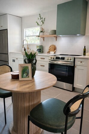 A modern kitchen with white cabinets, a round wooden table, two green upholstered chairs, a built-in oven, and a coffee machine. - Newly renovated duplex conveniently located between Vilano and downtown. (St. Augustine)