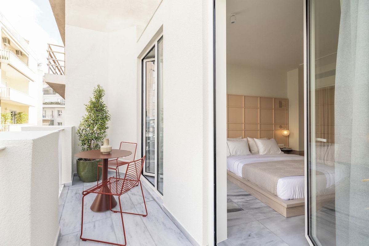 A balcony with a table and chairs, a potted plant, and a view of a bedroom with a bed and nightstand.