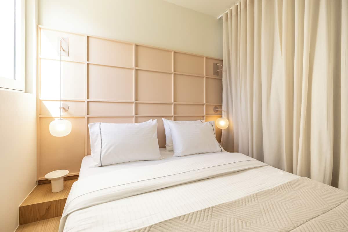 Una cama bien tendida con un cabecero, una mesita de noche con una lámpara y una ventana con cortinas.