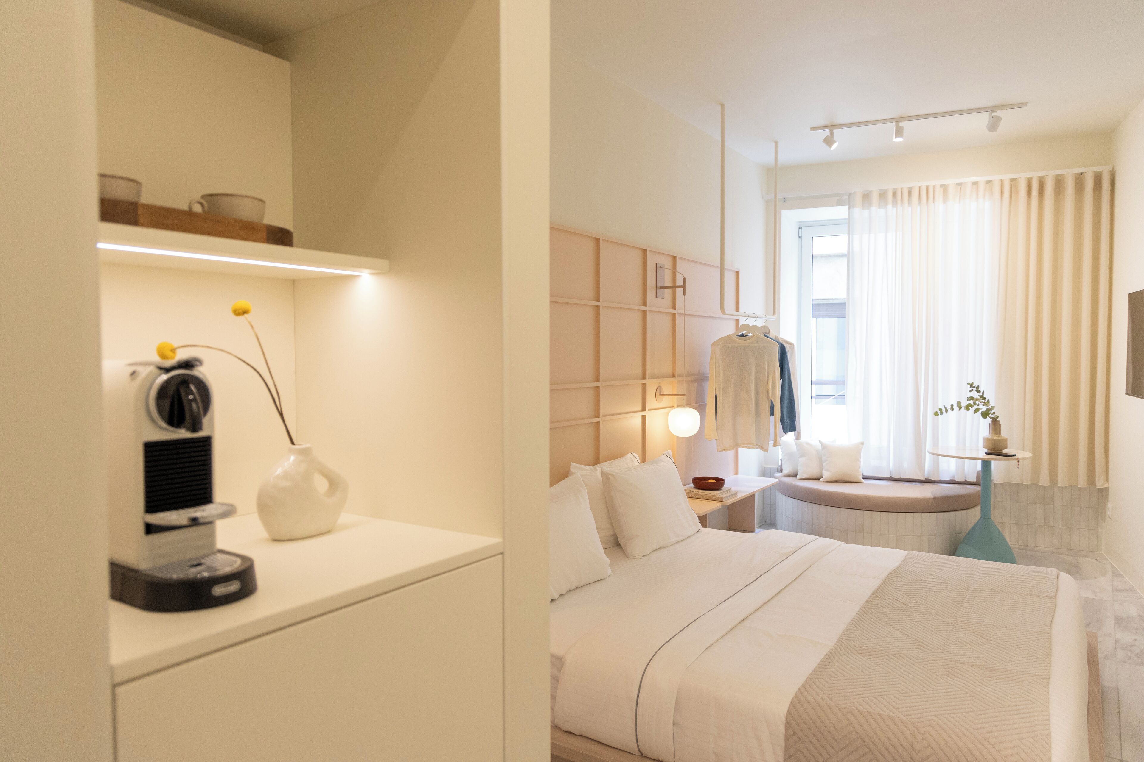 A modern hotel room with a neatly made bed, a bedside table with a lamp, a small seating area with a bench, and a window with curtains.
