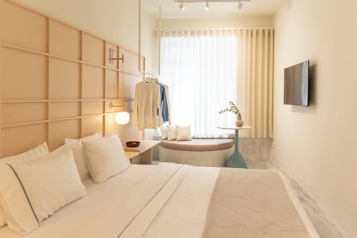 Habitación de hotel con una cama grande, una mesita de noche con lámpara, un televisor montado en la pared, una ventana con cortinas y una pequeña mesa redonda con una planta.