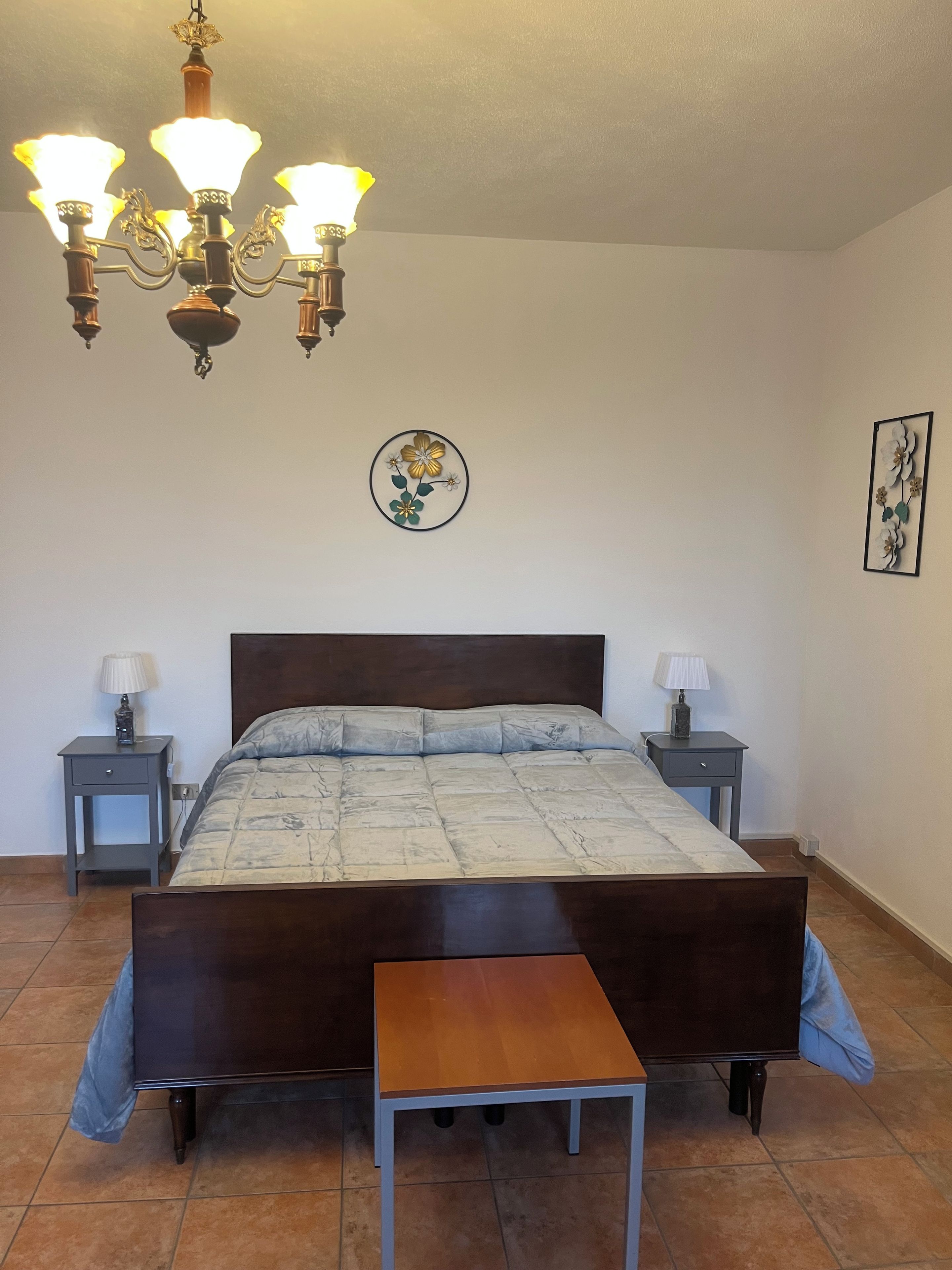 Un dormitorio con una cama grande, dos mesitas de noche con lámparas, una araña y un adorno floral en la pared.