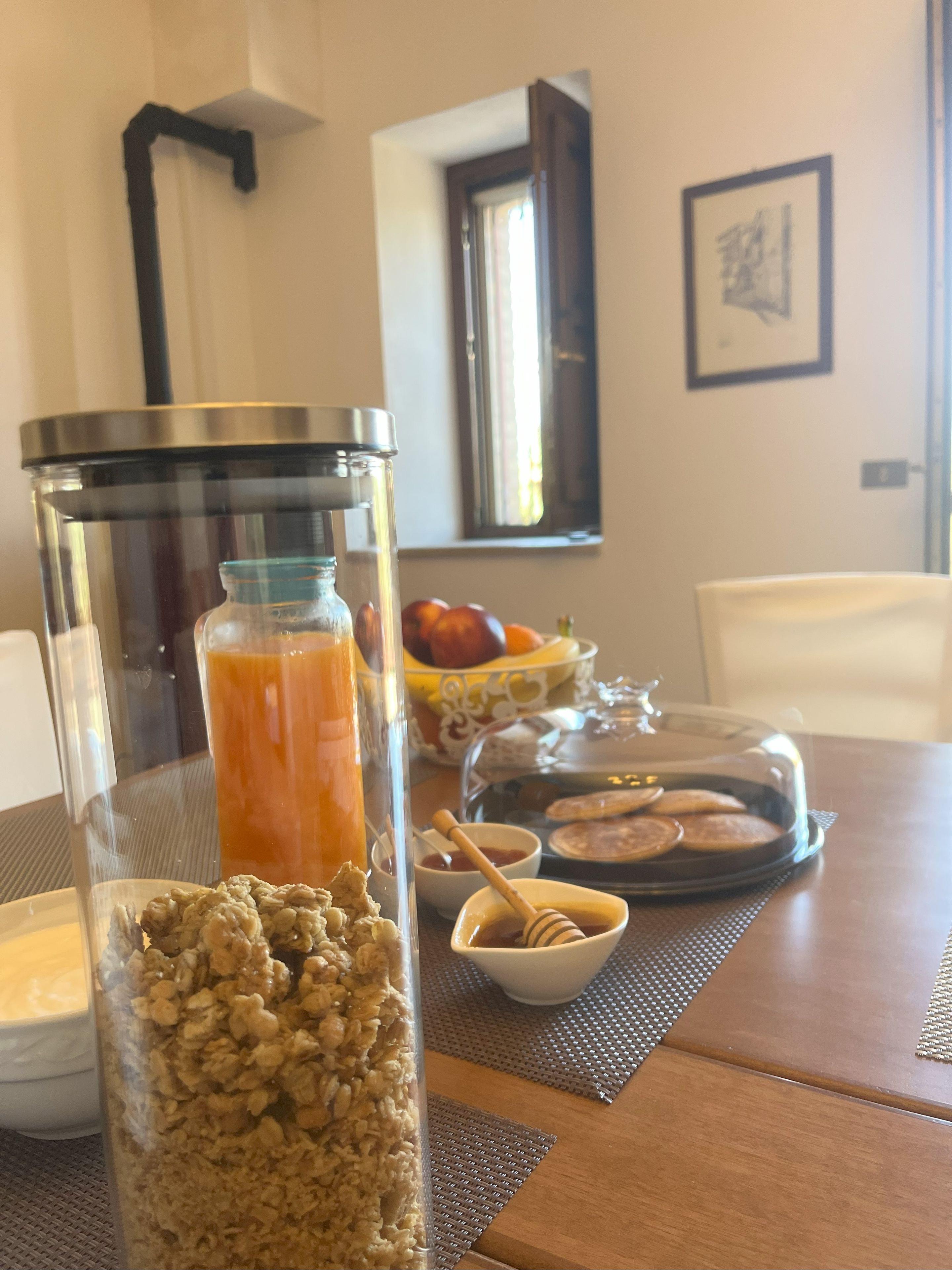 A breakfast table with a glass jar of granola, a glass bottle of orange juice, a bowl of honey, and a plate of pancakes.
