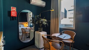 Et hotelværelse med en seng, et rundt bord med stole, et spejl, en stueplante og et vindue med gardiner.