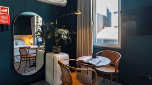 Et kompakt hotelværelse med en seng, et lille bord med en bog og en kop, to fletstole, et rundt spejl og et vindue med gardiner.
