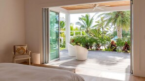 Un dormitorio con una cama grande, una silla con diseño de pez y vistas a una terraza de madera con palmeras y un edificio blanco.