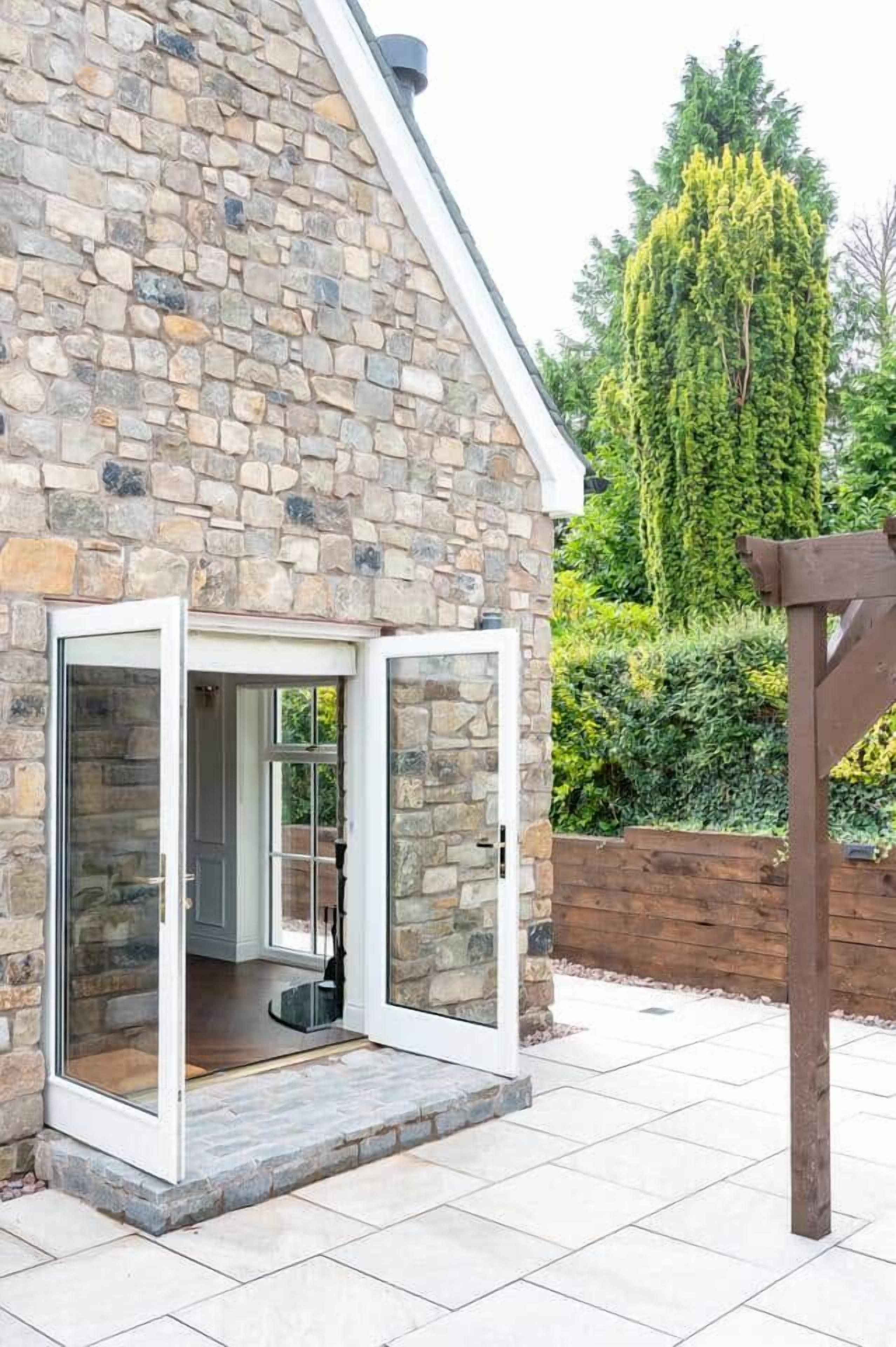 Un bâtiment en pierre avec une entrée dotée d’une porte en verre, une pergola en bois et un jardin bien entretenu.