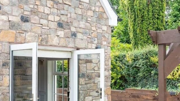A stone building with a glass door entrance, a wooden pergola, and a well-maintained garden.