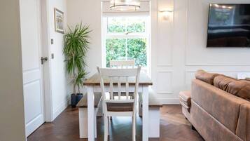 A dining area with a wooden table and chairs, a potted plant, a window with a view of greenery, a white wall-mounted TV, and a brown leather couch.