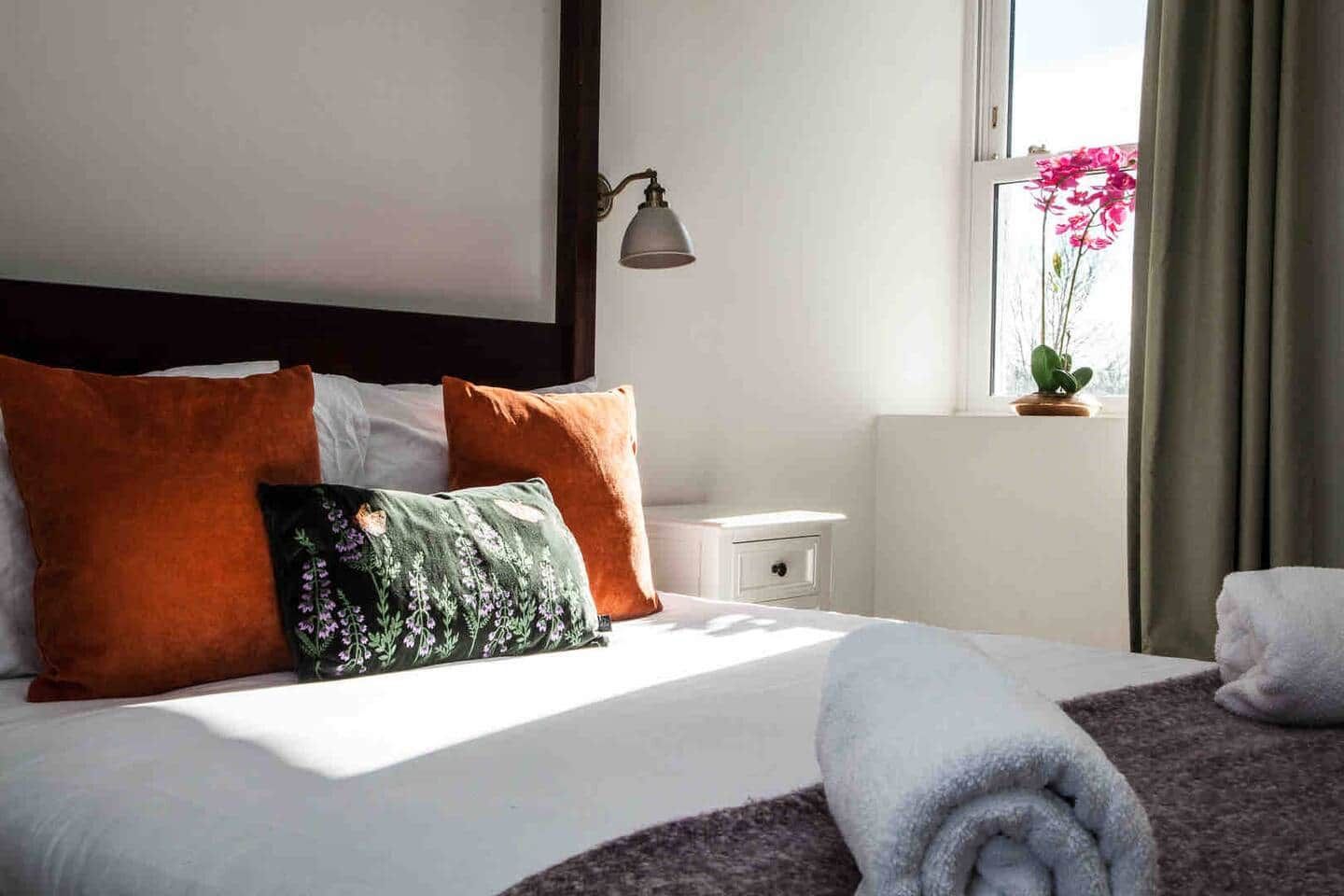 Ein ordentlich bezogenes Bett mit Kissen, ein Nachttisch, eine Lampe und ein Fenster mit Blick auf Blumen.