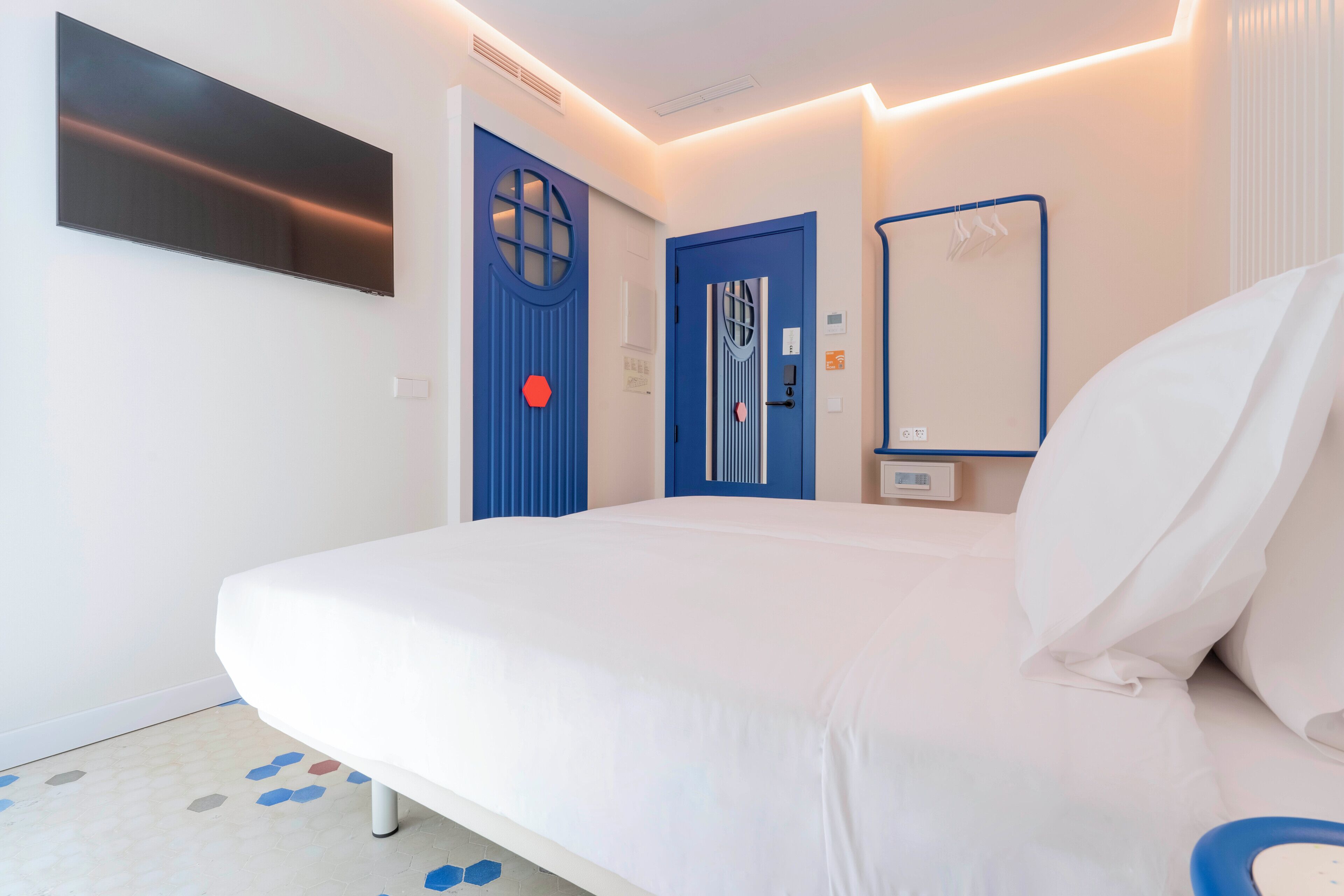A hotel room with a bed, two doors, a mirror, and a flat-screen TV mounted on the wall.