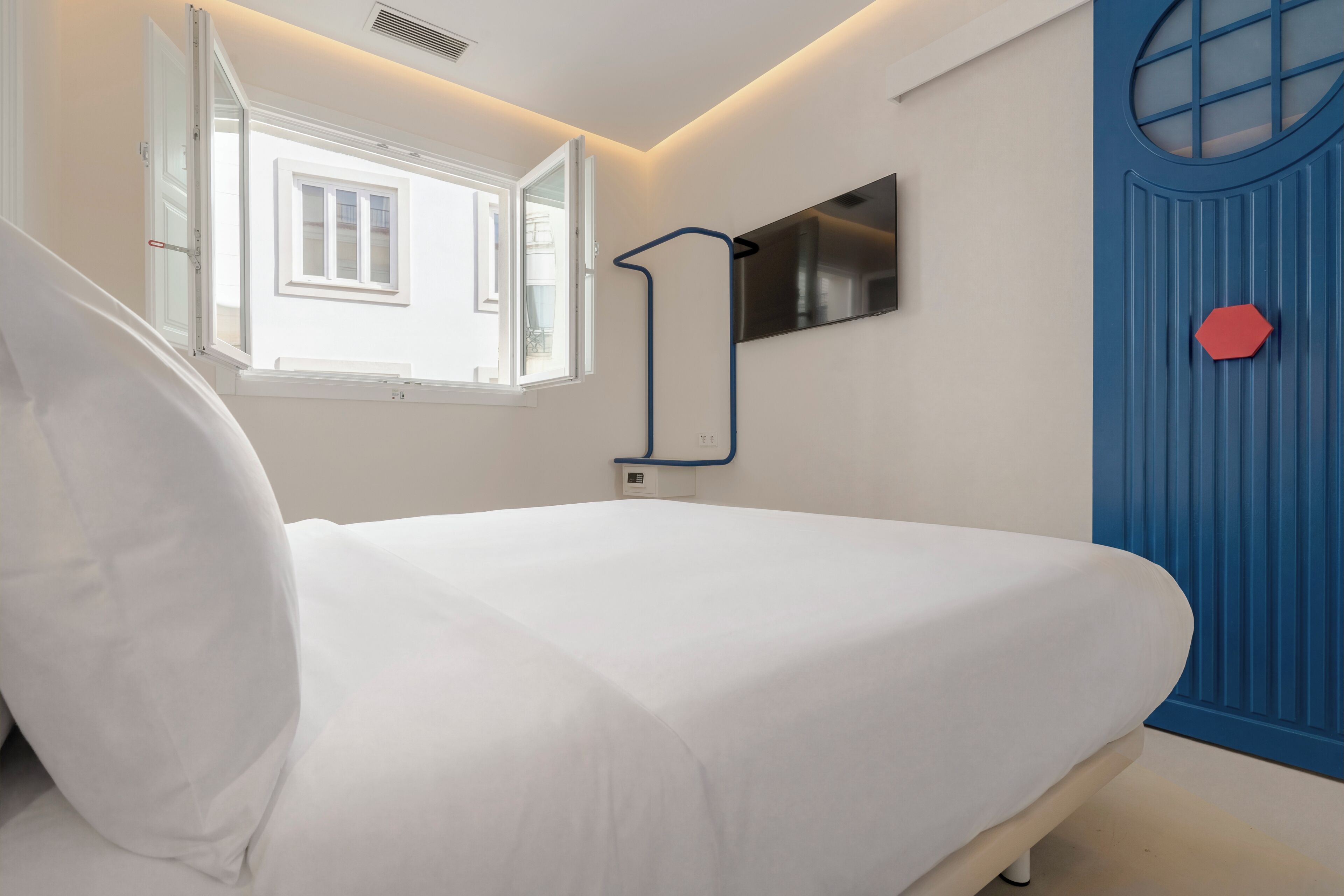 Una cama en una habitación con ventana, un televisor montado en la pared y una puerta azul con un objeto rojo.