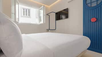 Una cama en una habitación con ventana, un televisor montado en la pared y una puerta azul con un objeto rojo.