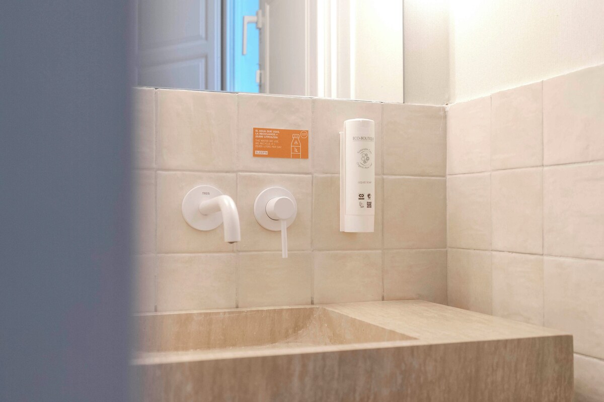 Un baño con pared de azulejos color beige, un dispensador de toallas blanco y una botella blanca en la pared.
