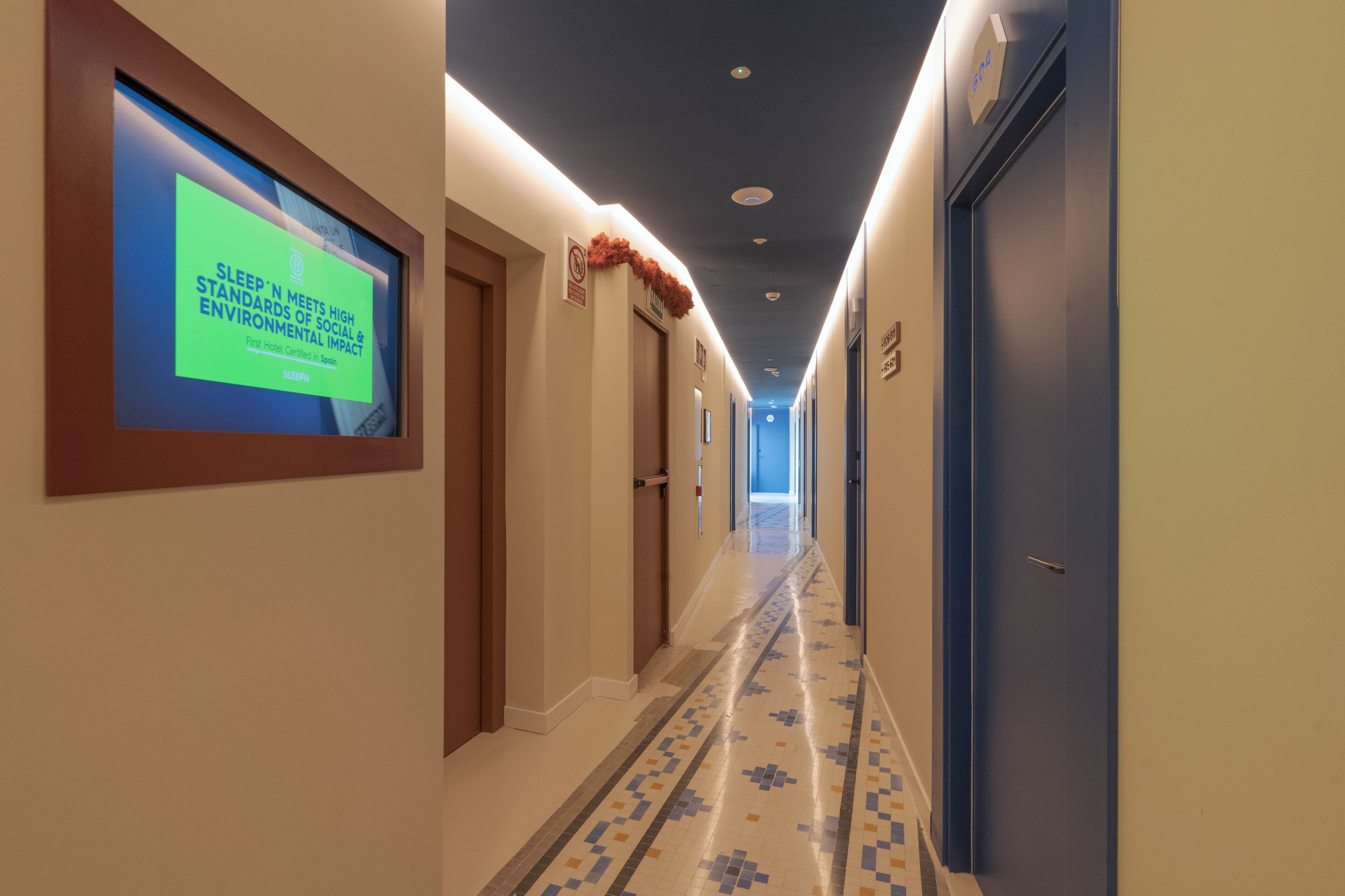 Un couloir d’hôtel avec un écran numérique sur le côté gauche, un sol à motifs et des portes de part et d’autre.