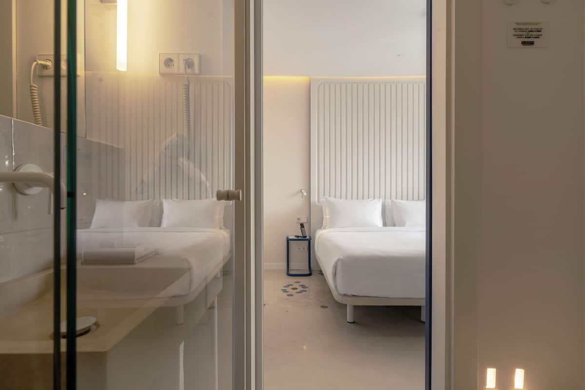 Habitación de hotel con dos camas, una mesita de noche, una pequeña mesa, un espejo y un radiador.