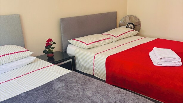 Quarto de hotel com duas camas, cada uma com colcha listrada em vermelho e branco, cabeceira cinza e uma mesinha com um ventilador e uma planta em vaso.