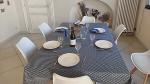 Een eettafel gedekt met borden, glazen en een fles, omringd door witte stoelen in een kamer met een betegelde vloer en een rode schildering aan de muur.
