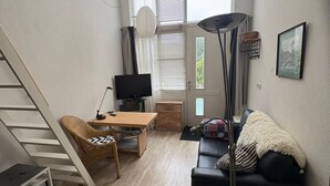 Een ruimtelijk ingerichte leefruimte met een houten vloer, een zwarte bank, een houten eettafel, een televisie, een trap en een groot raam met zonwering.
