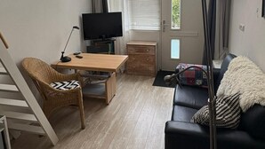 Een woonkamer met een houten vloer, een zwarte bank, een houten tafel, een televisie, een lamp en een raam met zonwering.
