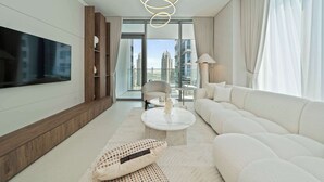 Ein modernes Wohnzimmer mit großem Flachbildfernseher, einem weißen Sofa, einem runden Couchtisch und Blick auf hohe Gebäude durch große Fenster.