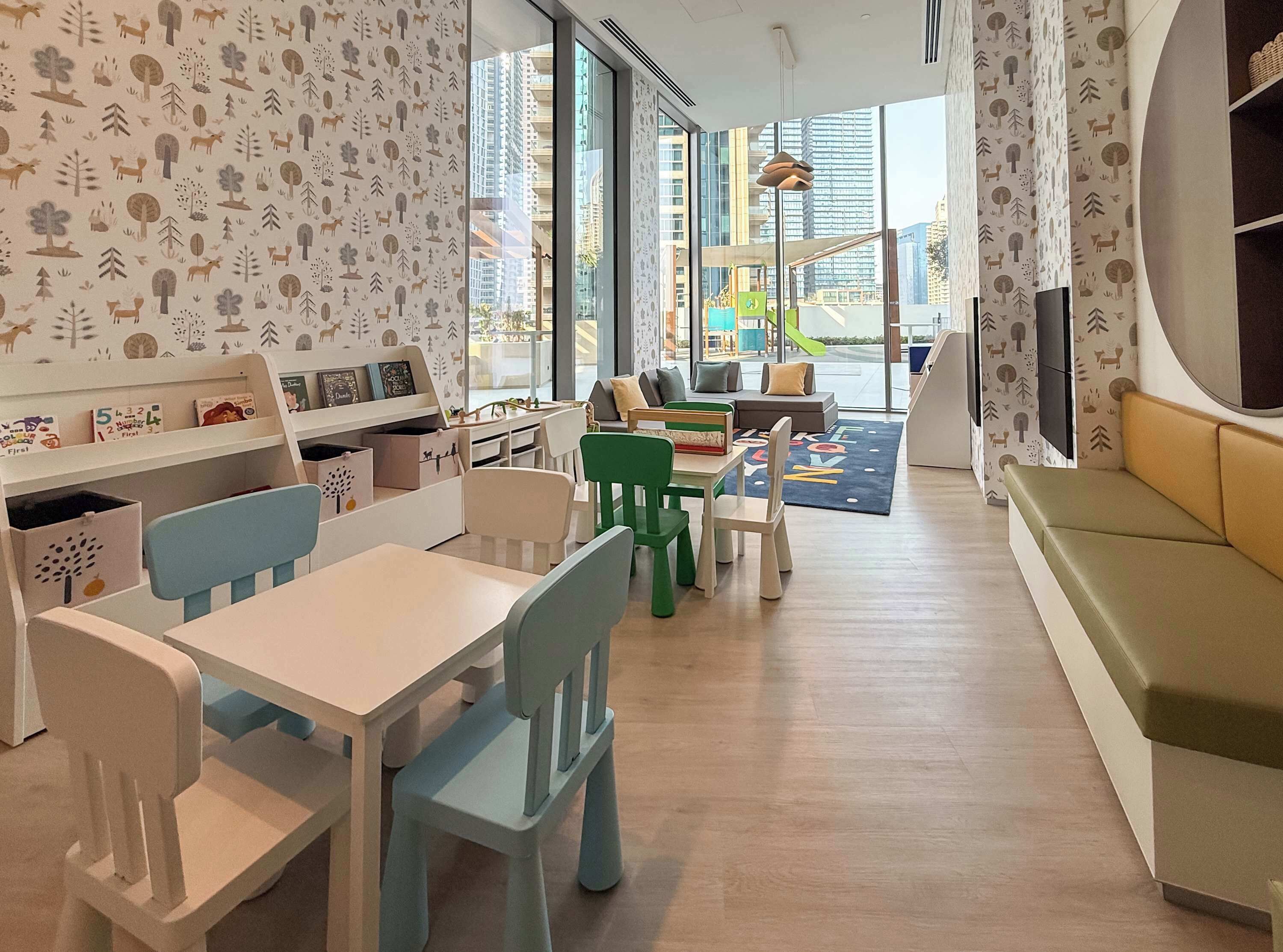 Un salón de juegos infantiles con sillas y mesas coloridas, y un ventanal con vistas a la ciudad.