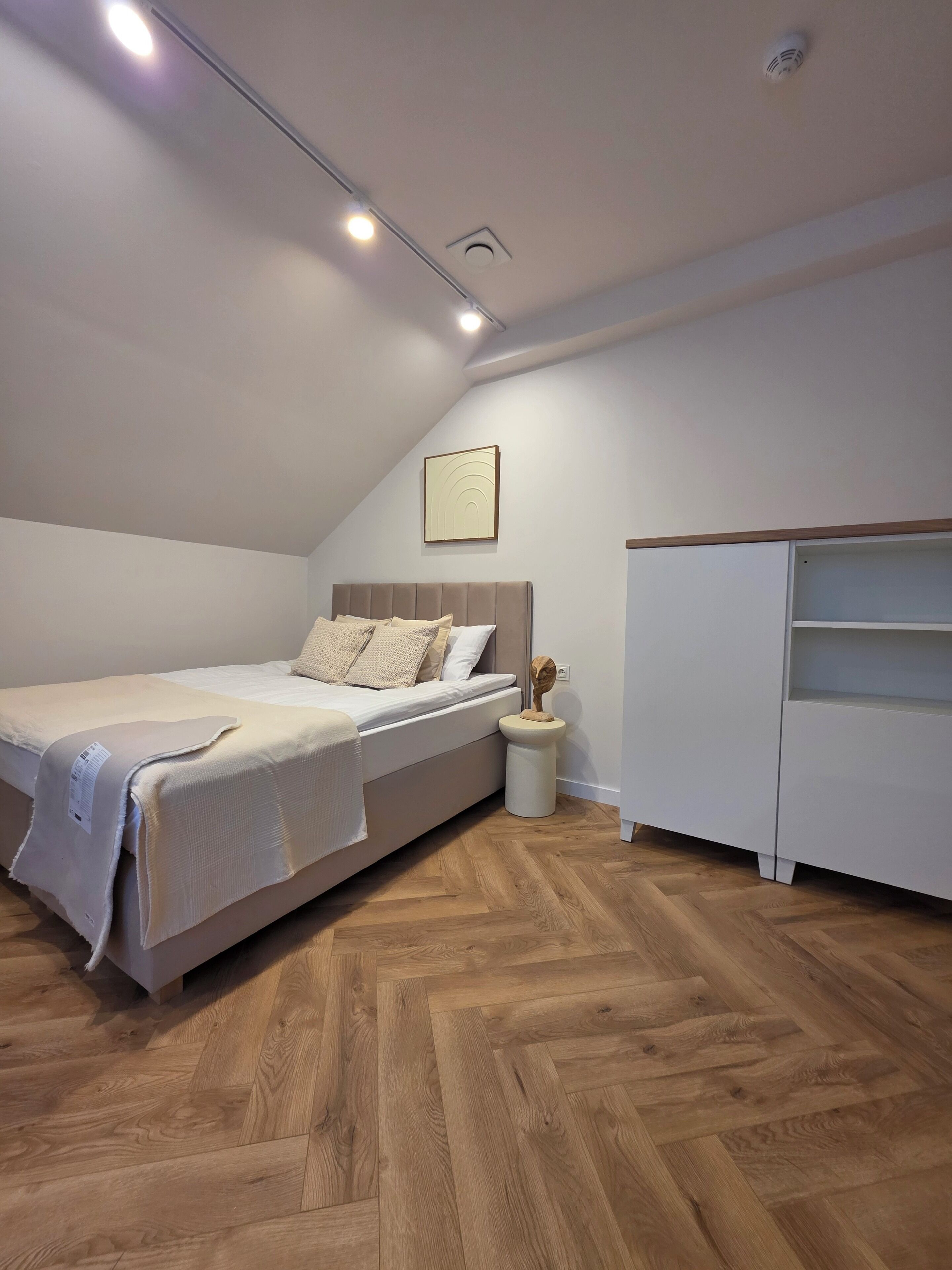 A bedroom with a bed, a nightstand, a white wardrobe, and a herringbone-patterned wooden floor.