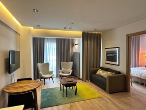 Une chambre d’hôtel équipée d’un téléviseur à écran plat, de deux fauteuils, d’un canapé, d’une petite table, d’un tapis vert, d’un lit avec une couette blanche et d’un tableau encadré au mur.