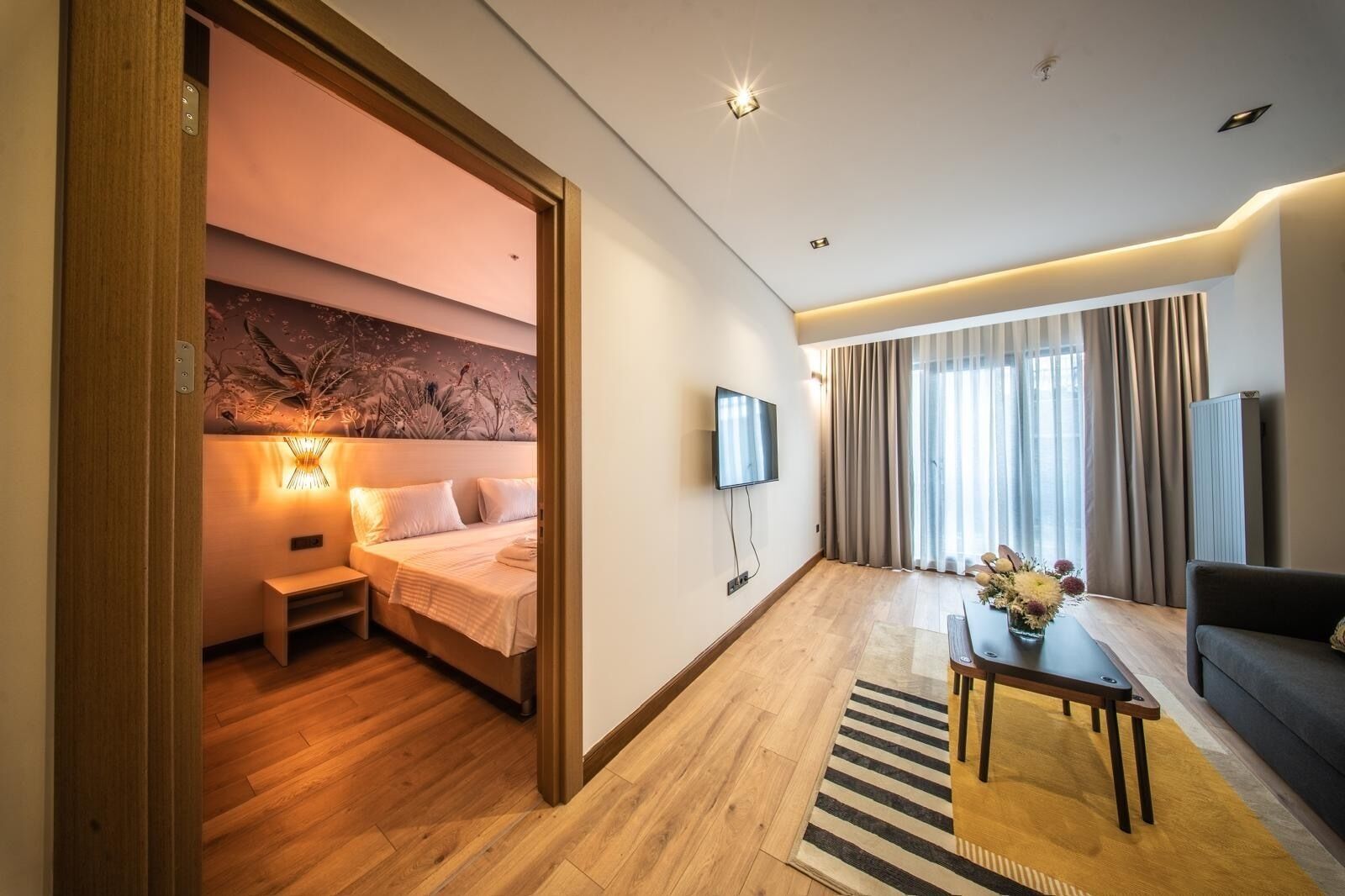 Een hotelkamer met een bed, een televisie, een bank, een kleine tafel met een vaas, en een raam met gordijnen.