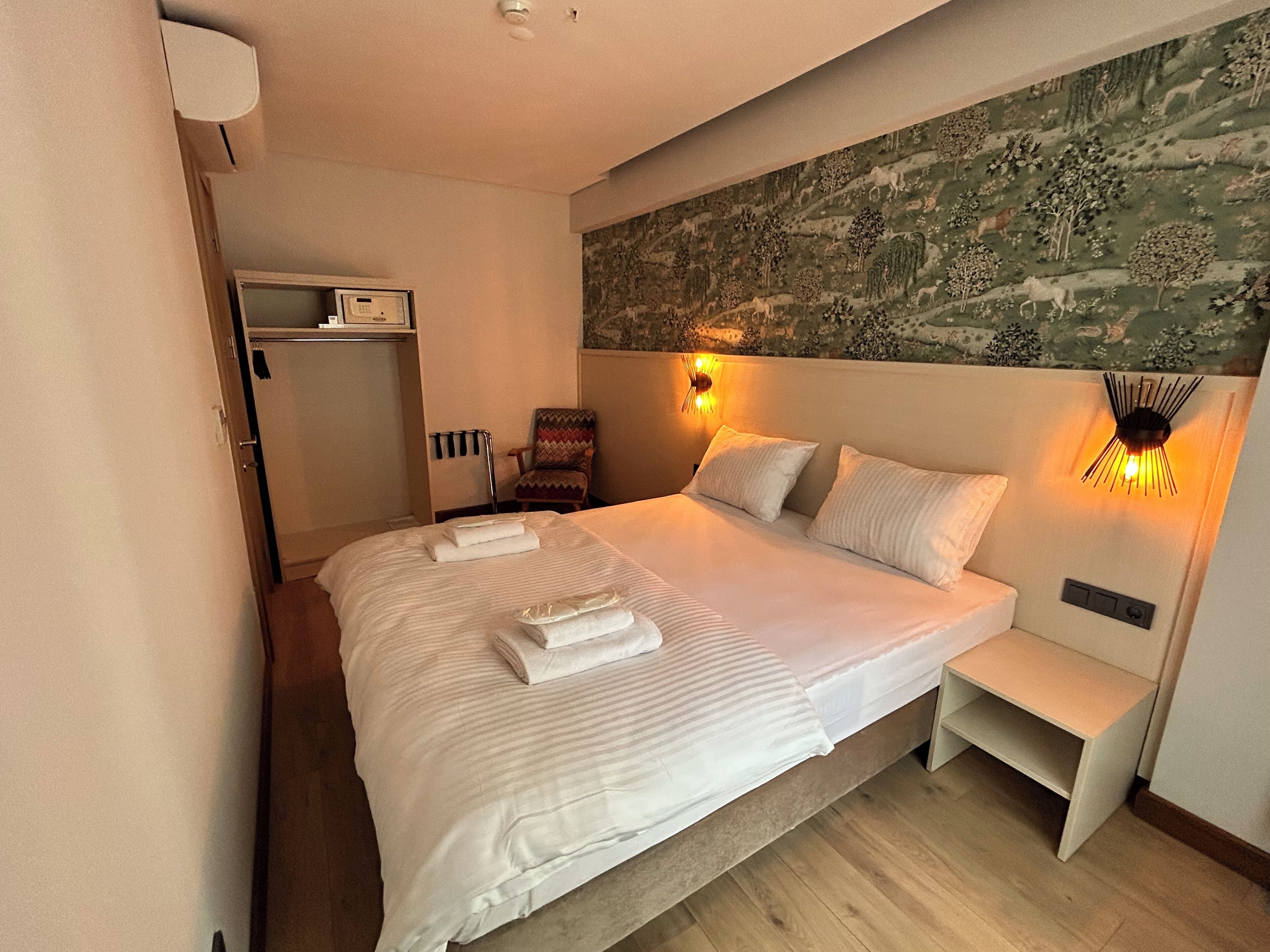 A neatly made bed with towels, a chair, a bedside table, and a wall with a nature-themed mural.