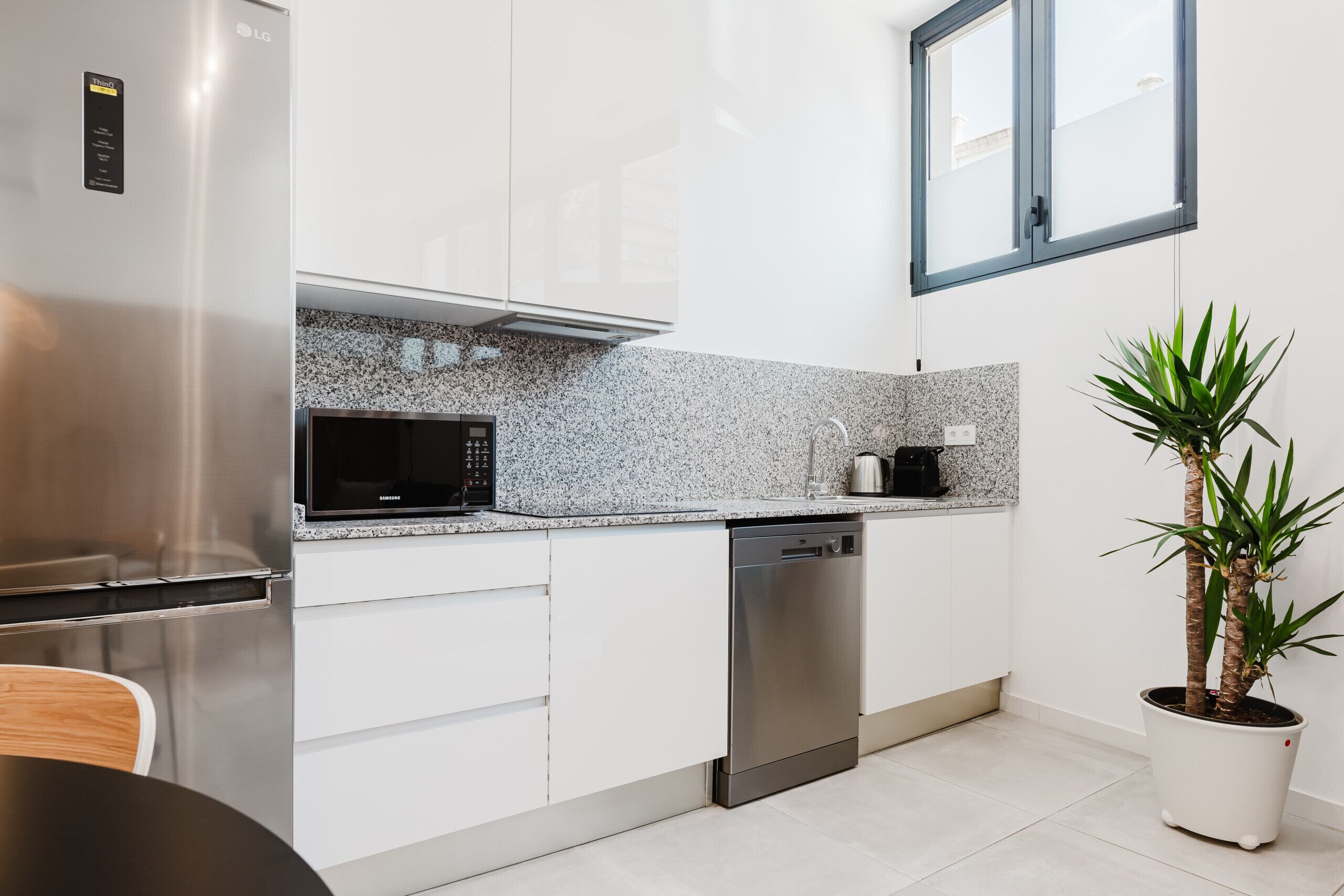 A modern kitchen with white cabinetry, a stainless steel refrigerator, a microwave, and a dishwasher. There is a window above the sink and a potted plant on the right.