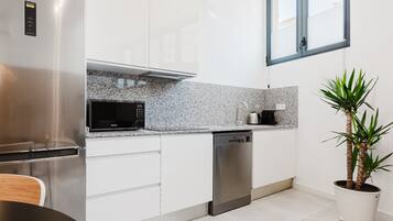 A modern kitchen with white cabinetry, a stainless steel refrigerator, a microwave, and a dishwasher. There is a window above the sink and a potted plant on the right.
