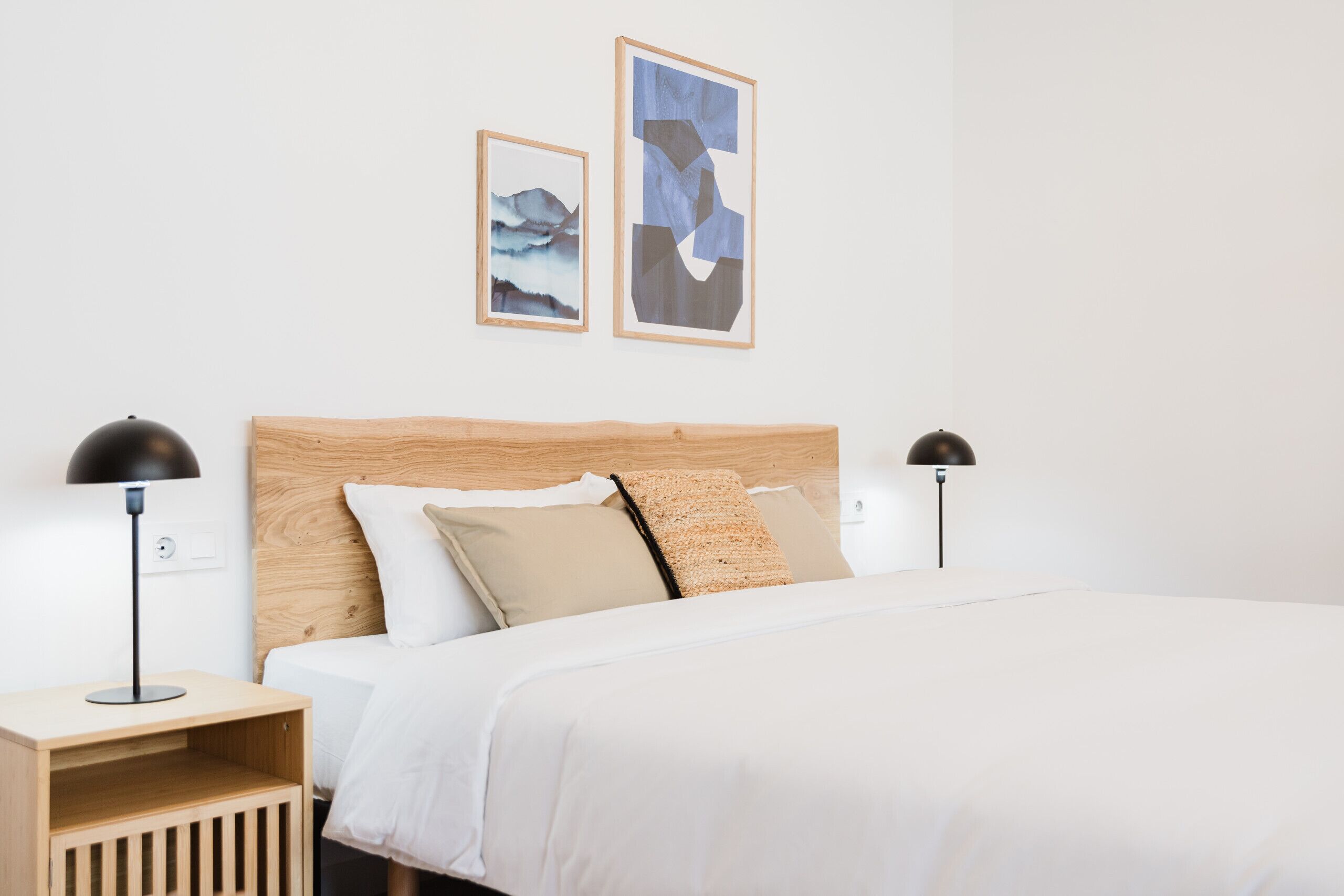 A neatly made bed with a wooden headboard, two bedside lamps, and two framed artworks on the wall.