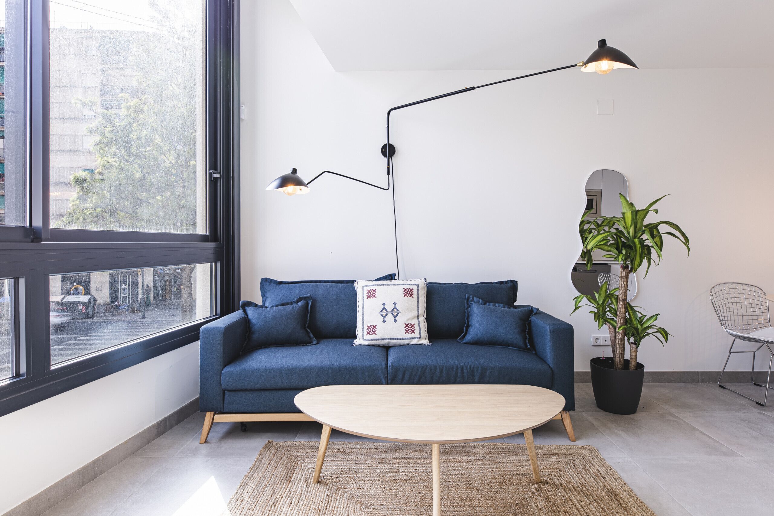 A modern living room with a sofa, a coffee table, a floor lamp, a potted plant, and a view through a large window.