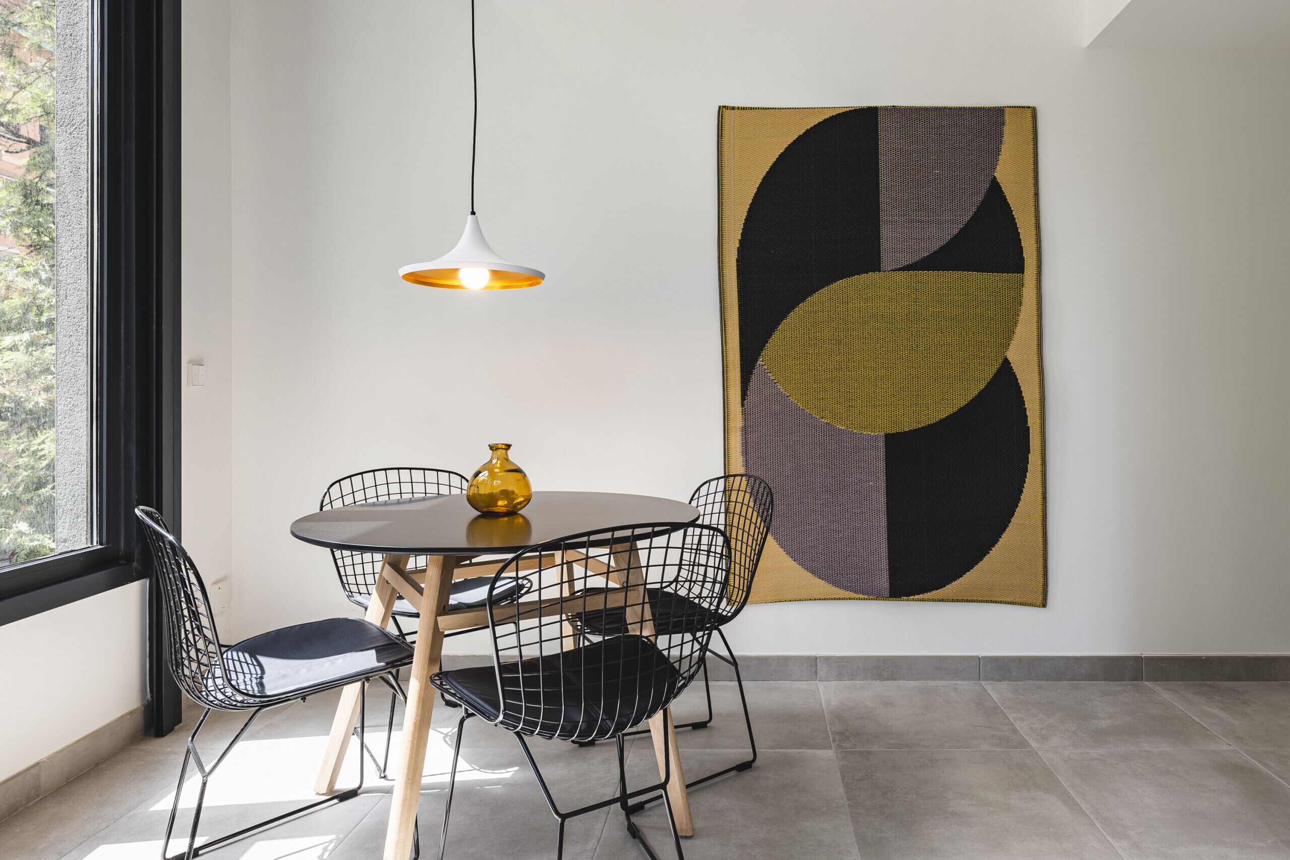Un comedor con una mesa redonda, sillas negras de estructura de alambre, un florero amarillo y una lámpara colgante.