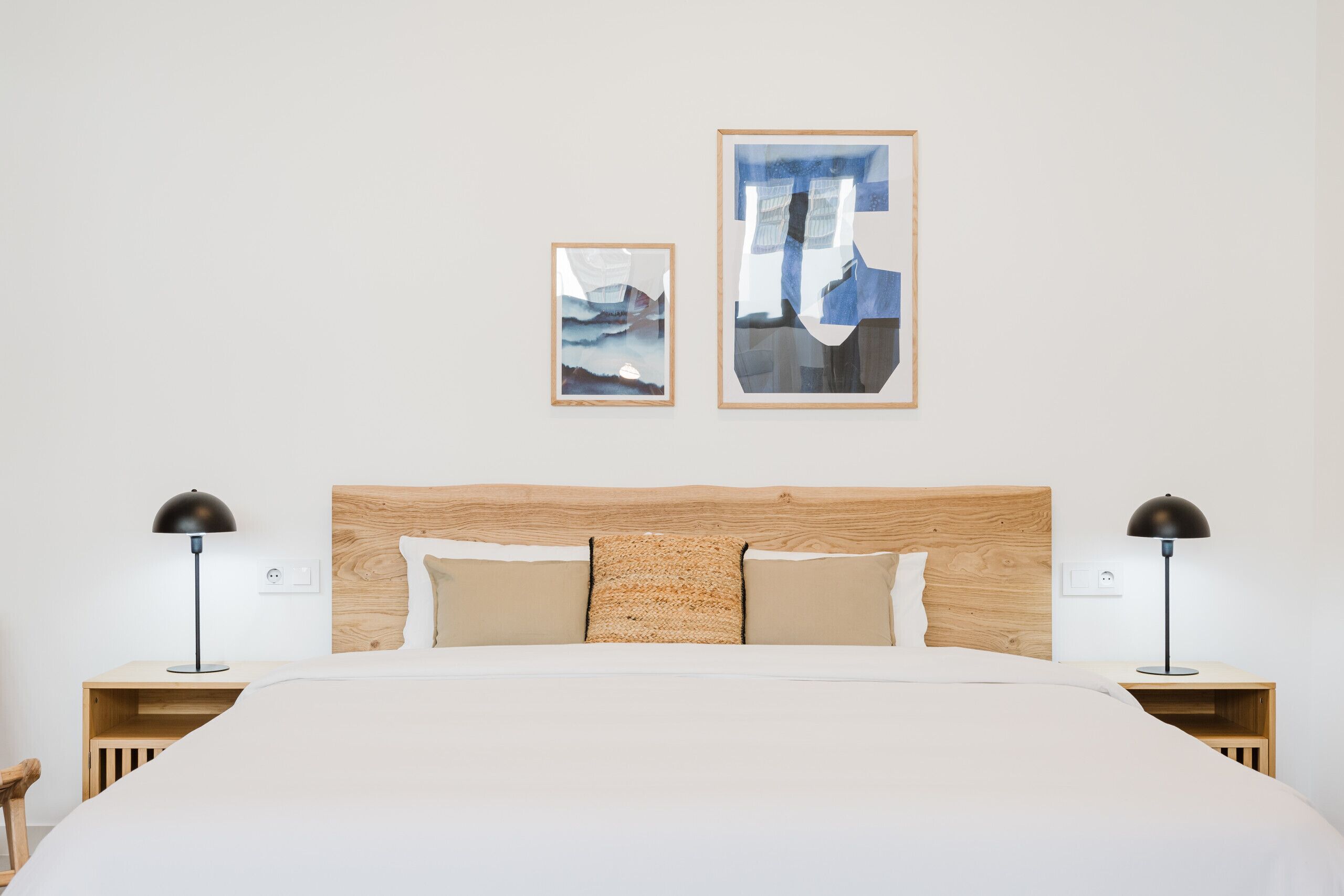 Una cama bien hecha con cabecera de madera, dos lámparas de noche y dos cuadros abstractos enmarcados en la pared.