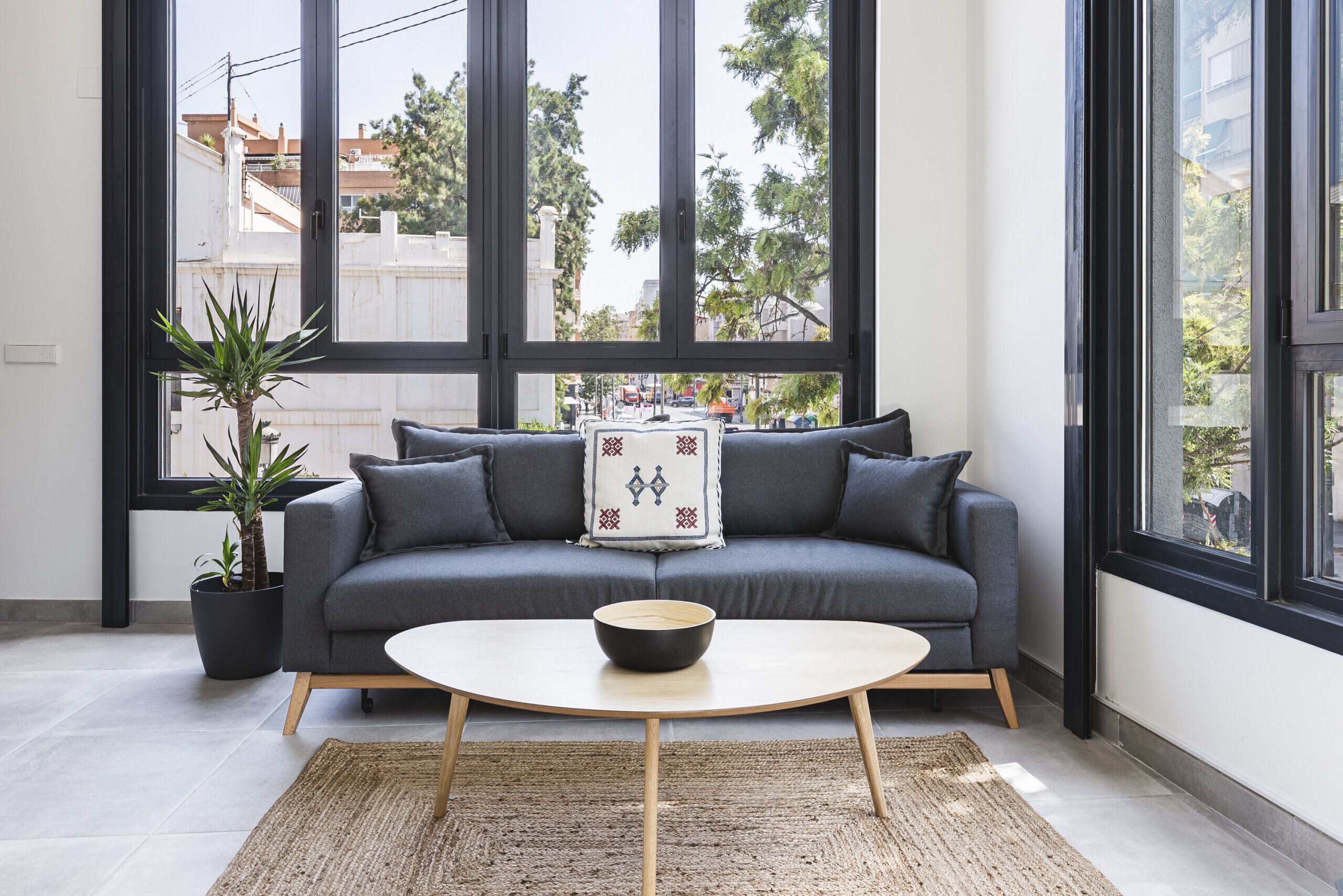 Un salon moderne avec un canapé gris, une table basse ronde, une plante en pot et de grandes fenêtres.