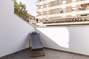 Un balcon avec une chaise en bois, un sol carrelé et, au fond, un bâtiment avec un toit recouvert de tuiles.