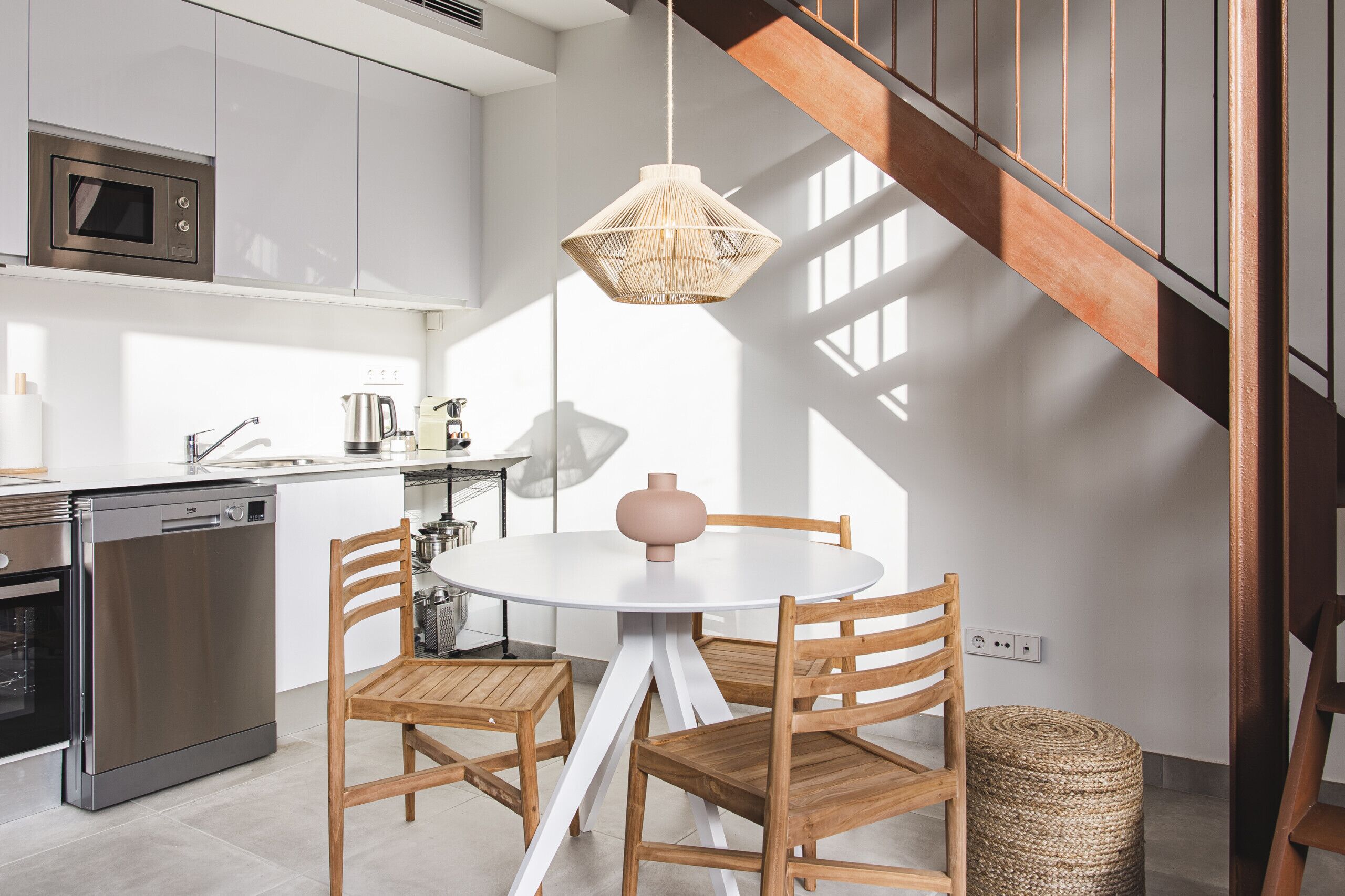 Una cocina moderna con electrodomésticos de acero inoxidable, una mesa redonda para comer con sillas de madera y una lámpara colgante de fibras naturales.