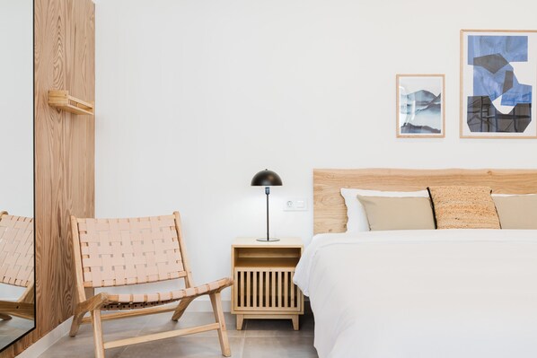 Une chambre avec une tête de lit en bois, une chaise en osier, une table de chevet avec une lampe, et deux œuvres d’art encadrées accrochées au mur.