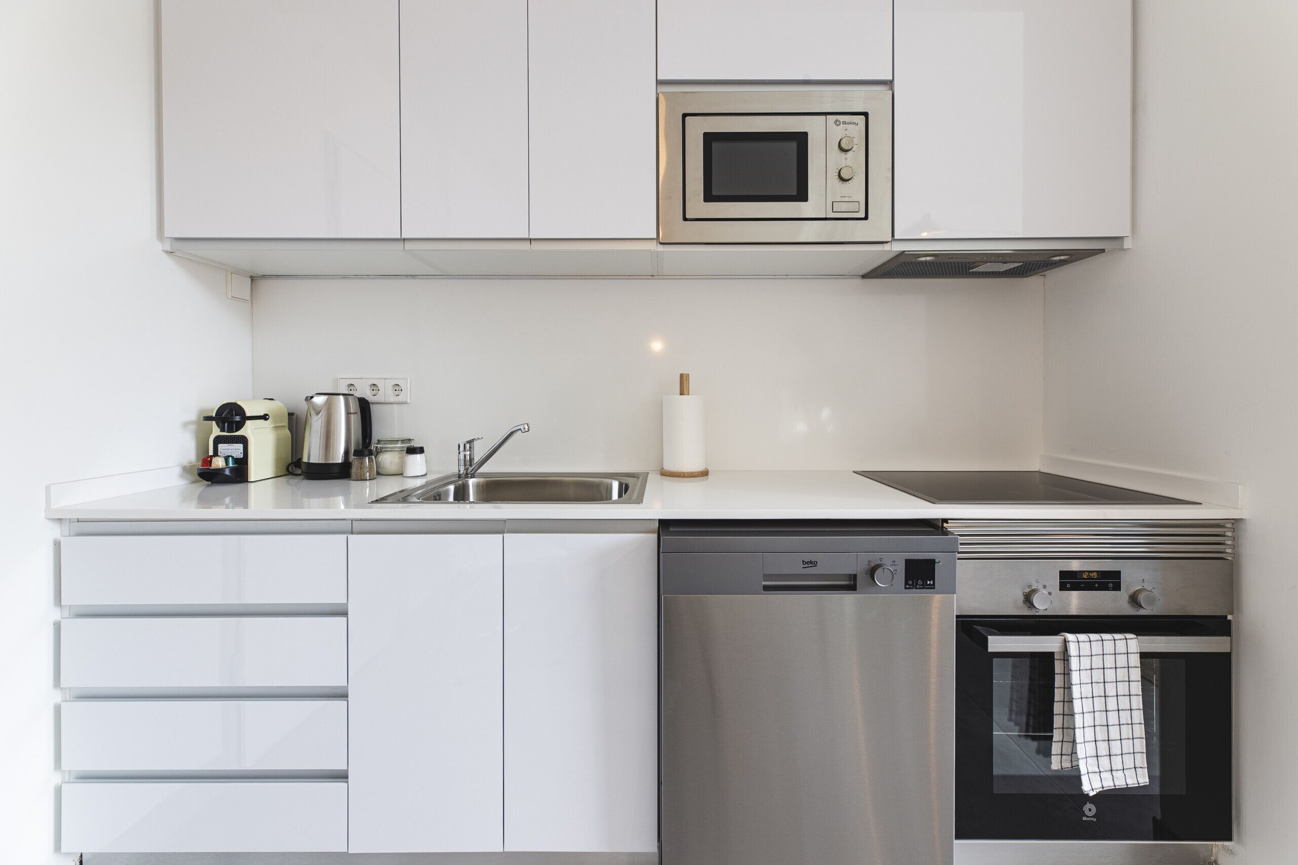 Una cocina moderna con armarios blancos, lavavajillas de acero inoxidable, horno empotrado y microondas.