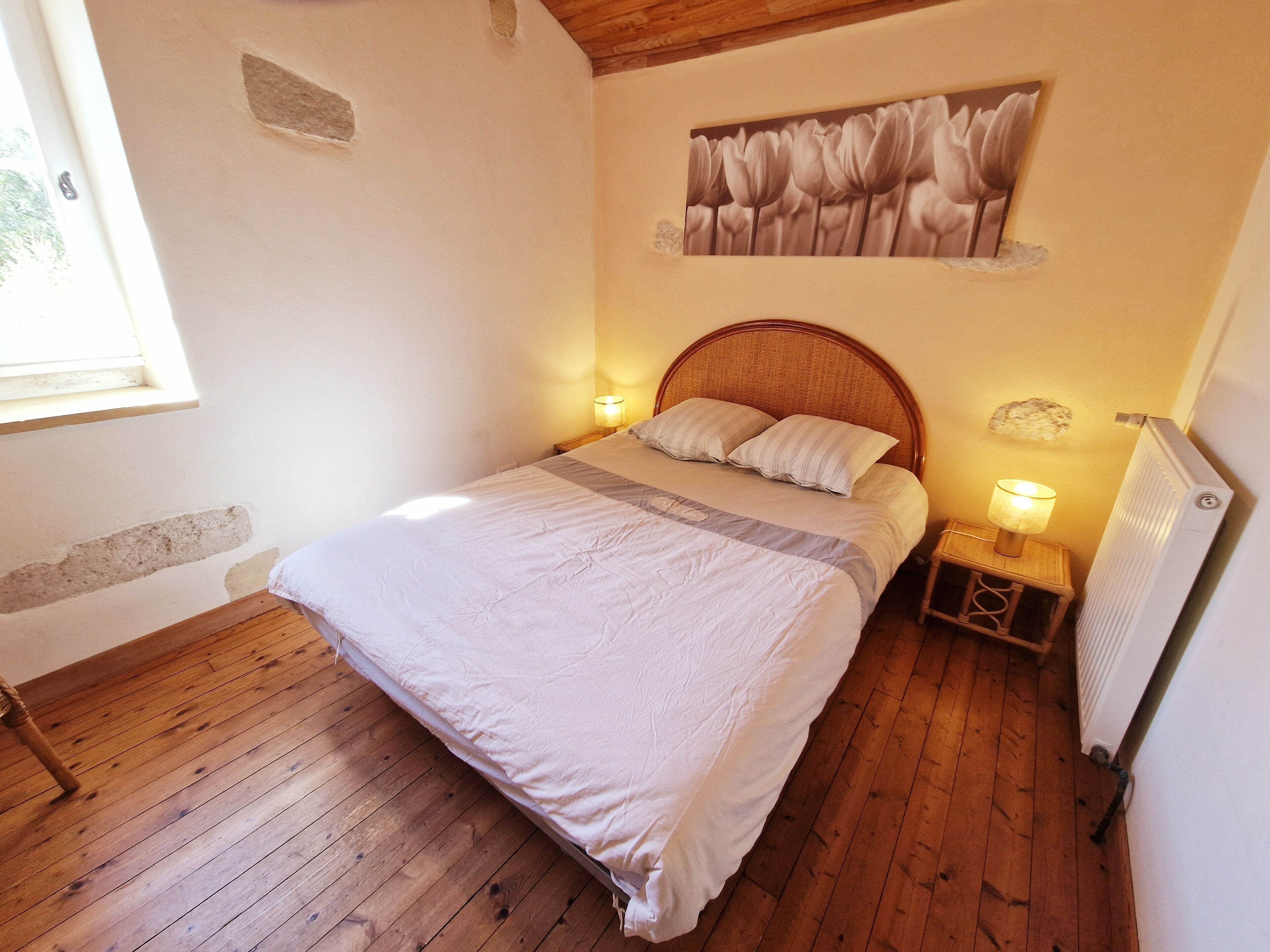 Quarto com chão de madeira, uma cama com cabeceira, duas luminárias de apoio, um radiador e uma janela.