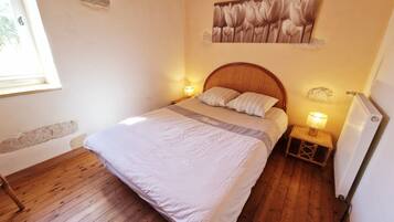 Quarto com chão de madeira, uma cama com cabeceira, duas luminárias de apoio, um radiador e uma janela.