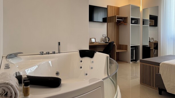 Een moderne hotelkamer met een groot ligbad, een bureau met stoel, een flatscreen televisie aan de wand en een inloopkast met planken.