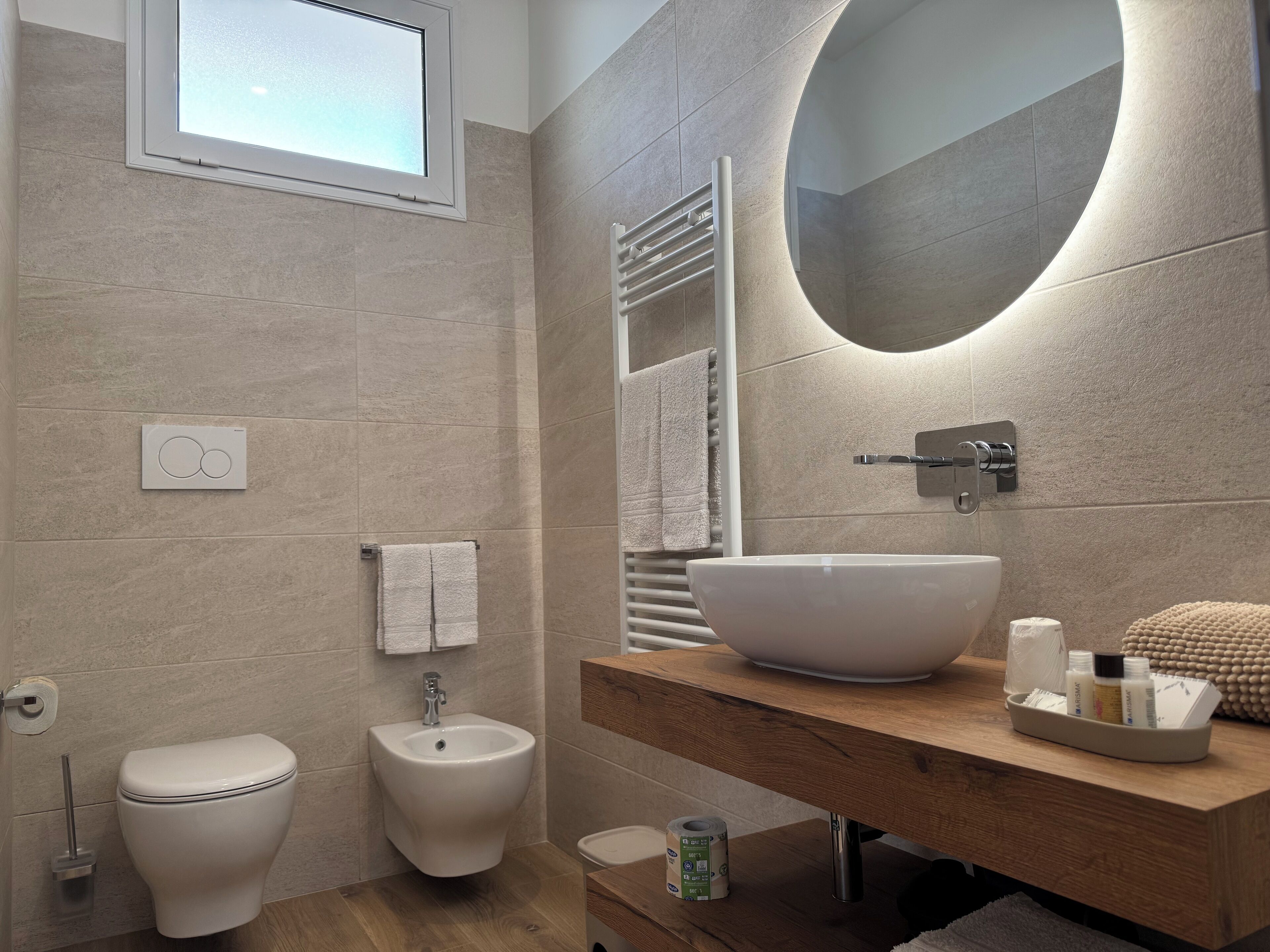 A modern bathroom with a bidet, a large oval sink, and a mirror with integrated lighting.