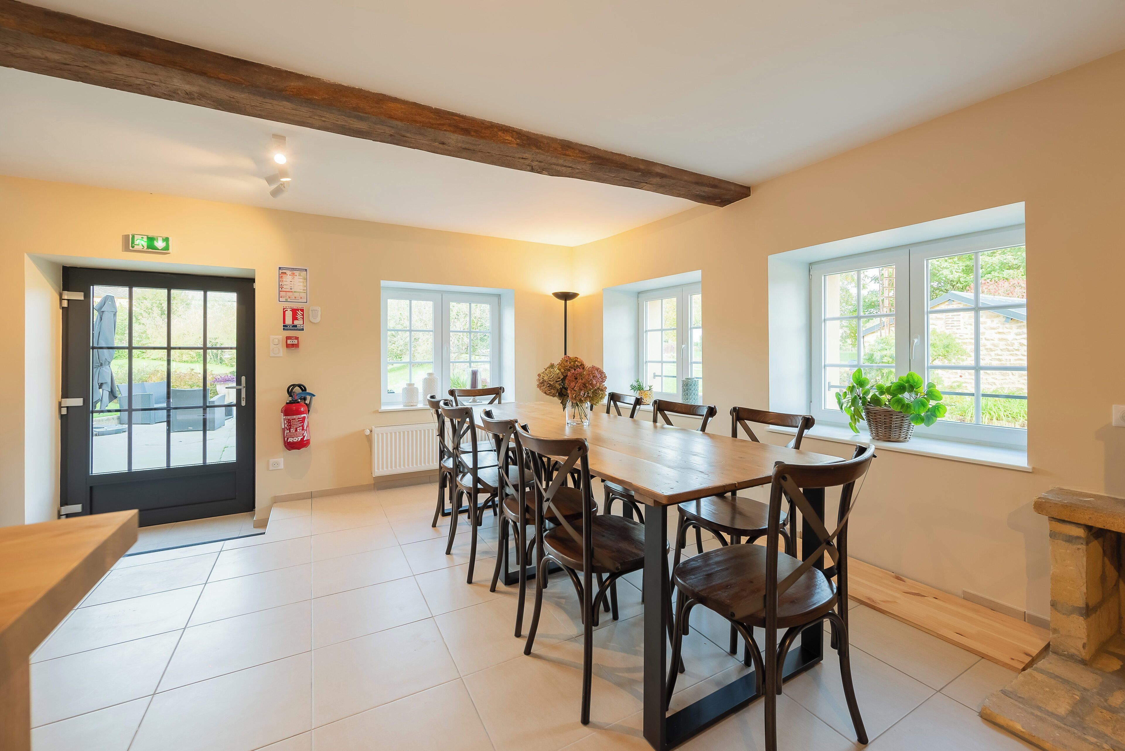A dining area with a long wooden table and chairs, a fire extinguisher, and a window with a plant.