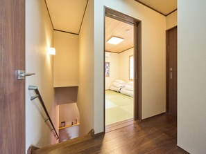 Um corredor com piso de madeira, uma porta de madeira e uma cama com um colchonete verde no quarto adjacente.
