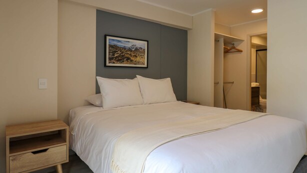 Una cama bien hecha con sábanas blancas en una habitación con una pared de acento gris y un cuadro de paisaje enmarcado.